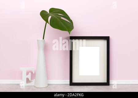 Modèle de maquette élégant avec cadre photo vertical en bois noir avec lettre E en bois mat et feuille de Monstera deliciosa dans un vase devant un mur rose Banque D'Images