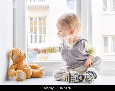 Enfant en quarantaine à la maison à la fenêtre mettant un masque médical sur son ours en peluche malade, pour la protection contre les virus pendant le coronavirus COVID-19 et f Banque D'Images