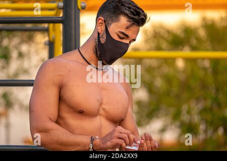 Jeune homme Latino sans chemise et pantalon noir et masque désinfectant ses mains avec du gel hydroalcoolique pendant la formation un parc de calisthéniques Banque D'Images