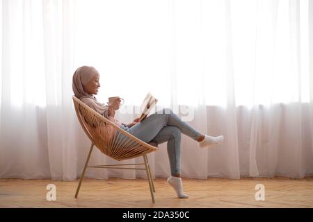 Vue latérale de la jeune femme noire dans hijab boire un café et lire dans un fauteuil confortable à l'intérieur, espace de copie Banque D'Images