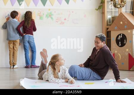 Portrait complet d'une enseignante souriante assise sur le sol avec des enfants qui dessinant des photos tout en appréciant la classe d'art à Noël, espace de copie Banque D'Images