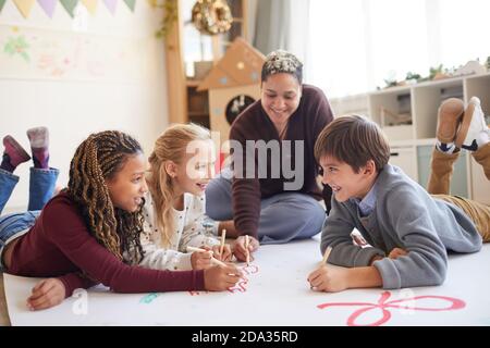 Portrait complet d'une enseignante souriante assise sur le sol avec un groupe d'enfants multiethnique qui dessine des photos tout en profitant de la classe artistique et de l'espace de copie Banque D'Images