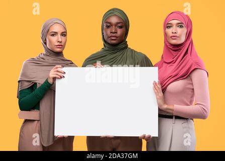 Trois femmes musulmanes graves tenant un tableau de papier vierge, fond jaune Banque D'Images