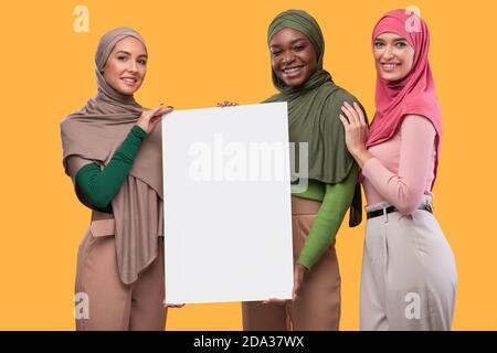 Trois musulmanes en hijab tenant une affiche vide, fond jaune Banque D'Images