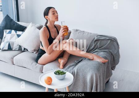 Une jeune femme au corps mince dans des vêtements de sport s'assoit canapé et mange des aliments diététiques sains à l'intérieur à la maison Banque D'Images