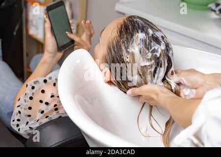 femme dans un salon de beauté. soins capillaires. coiffeur applique un masque aux cheveux du client. femme regardant son téléphone portable Banque D'Images