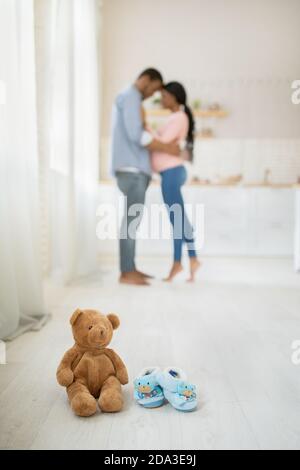 Ours en peluche et chaussons de bébé mignons sur le sol à l'intérieur et couple enceinte noire qui se hale sur l'arrière-plan, mise au point sélective Banque D'Images