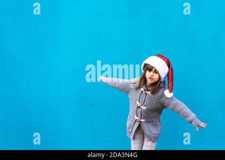 Jolie petite fille portant le chapeau du père noël volant avec ouvert Bras comme un avion - drôle de gamin jouant contre un Fond de mur bleu à l'extérieur à Noël t Banque D'Images