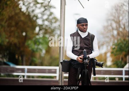 Jeune photographe afro-américain professionnel tenant un appareil photo professionnel avec un équipement professionnel. Afro cameraman portant le duraq noir faire des vidéos. Banque D'Images