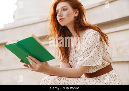 Image de la petite fille de gingembre concentrée dans un livre de lecture de chapeau de paille assis dans les escaliers le jour de l'été Banque D'Images