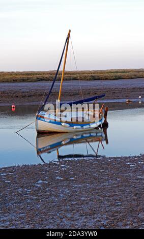 Petit bateau avec voile et réflexion amarré dans le port de North Norfolk à basse eau à Burnham Overy Staithe, Norfolk, Angleterre, Royaume-Uni. Banque D'Images