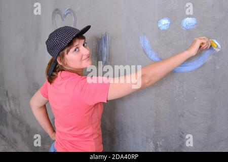 Une femme en casquette dessine un sourire sur le mur, gros plan. Le peintre peint le mur avec de la peinture blanche. Réparez vous-même la nouvelle maison. Banque D'Images