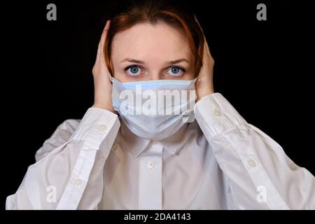 Une femme avec un regard effrayé dans un masque médical sur fond noir. La fille tient sa tête avec les deux mains dans la droite. Concept de personnes qui sont isola Banque D'Images
