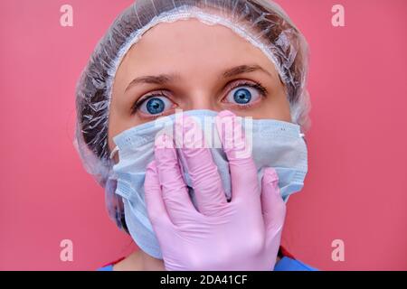 Portrait d'une femme médecin avec une droite dans les yeux, gros plan. Infirmière dans un uniforme bleu et un masque de protection sur fond rose. Banque D'Images