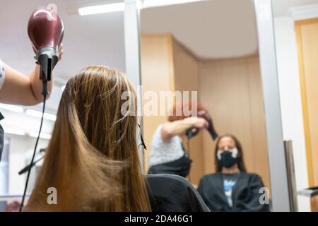 Coiffeur professionnel, styliste faisant la coiffure, le séchage et le soufflage des cheveux avec un sèche-cheveux dans un salon de beauté, réfléchi dans un miroir.portant un masque médical Banque D'Images