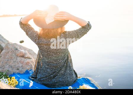 Une belle jeune fille rencontre l'aube lors d'un pique-nique avec un petit déjeuner romantique. La fille est assise face à la mer et au soleil, tenant son chapeau avec ses mains. Banque D'Images