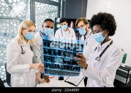 Équipe de médecins ou de scientifiques multiethniques, portant des masques médicaux de protection, regardant la tomodensitométrie du patient et discutant des résultats de Banque D'Images