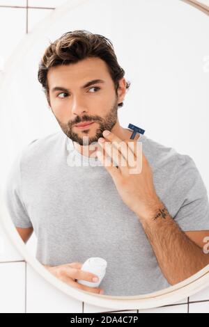 Homme barbu touchant le menton tout en tenant le rasoir et la mousse à raser près du miroir dans la salle de bains Banque D'Images