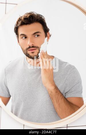 Homme barbu regardant le miroir tout en appliquant de la mousse de rasage salle de bains Banque D'Images