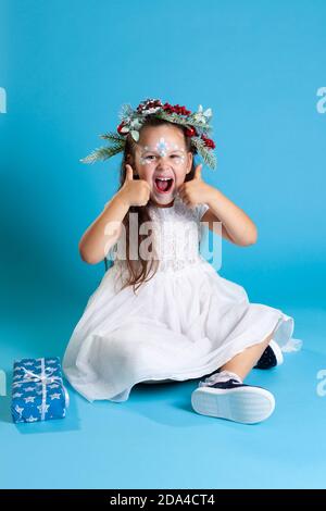 Joyeuses filles dans une robe de Noël blanche, portant une couronne et des baskets assises sur le sol et montrant deux pouces vers le haut, isolées sur un fond bleu Banque D'Images