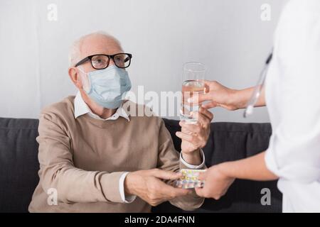 homme âgé dans le masque médical prenant un verre d'eau et pilules de l'infirmière gériatrique sur un premier plan flou Banque D'Images