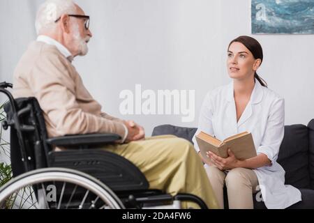 travailleur social en manteau blanc livre de lecture aux personnes âgées handicapées homme en fauteuil roulant sur un premier plan flou Banque D'Images