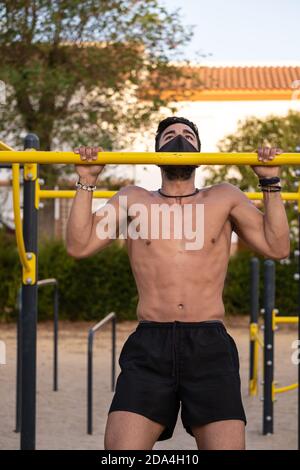 Jeune homme latino en pantalon noir et masque faisant des exercices dans un parc de calisthéniques sans t-shirt Banque D'Images