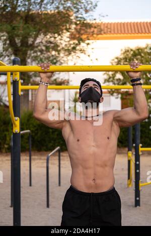 Jeune homme latino en pantalon noir et masque faisant des exercices dans un parc de calisthéniques sans t-shirt Banque D'Images