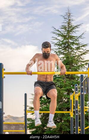 Latin jeune homme sans chemise et pantalon noir et masque faire des pull-ups sur un bar de parc de calisthéniques Banque D'Images