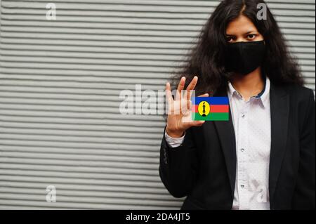 Femme asiatique en tenue officielle et masque de protection noir tenir le drapeau de la Nouvelle-Calédonie à portée de main sur fond gris. Coronavirus au concept de pays. Banque D'Images