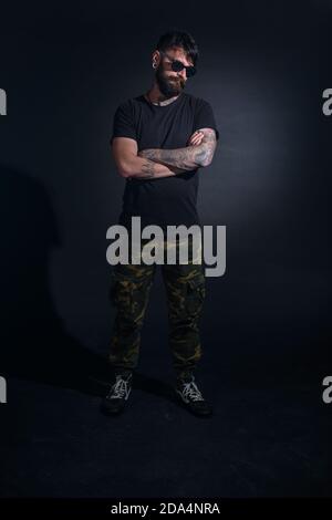 Modèle mâle barbu avec tatouages et bras croisés, vêtu d'un t-shirt noir et de lunettes de soleil pose sur fond noir. Banque D'Images