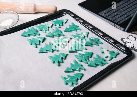 Cuisiner des biscuits de Noël selon une recette sur Internet. Ordinateur portable ouvert, plaque de cuisson avec biscuits sous forme d'arbres de Noël. Horizontale Banque D'Images