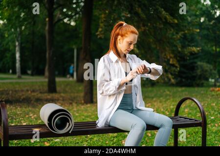 Photo moyenne d'une belle jeune femme aux cheveux rouges assise sur un banc dans un parc de la ville à l'aide de la montre intelligente. Banque D'Images