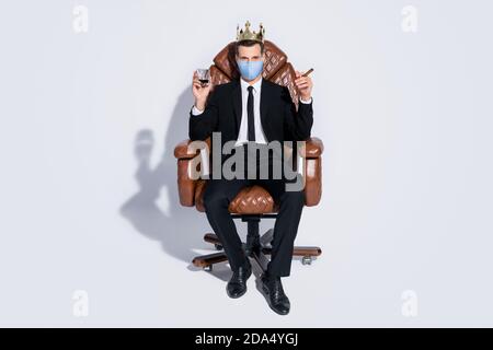 Photo du corps complet d'un beau homme d'affaires assis chaise de bureau tenez le verre à whisky cigare cubain couronne dorée porter tissu bleu masque facial gris isolé Banque D'Images