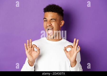 Un homme afro-américain effronté, s'assure de vous dans quelque chose, montrant des signes corrects dans l'approbation, se tenant heureux sur fond violet Banque D'Images