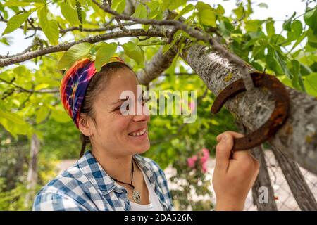 Jeune femme latine souriante avec foulard et chemise à carreaux bleue accrocher un fer à cheval rouillé sur une branche d'arbre Banque D'Images