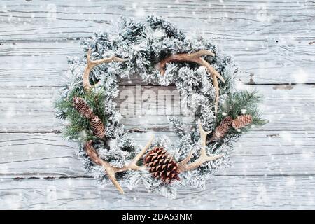 Campagne floquée ou chasseurs couronne de Noël sur fond de bois blanc avec neige tombant. Fabriqués avec des cônes de pin, des branches de sapin et des bois de cerf. Banque D'Images