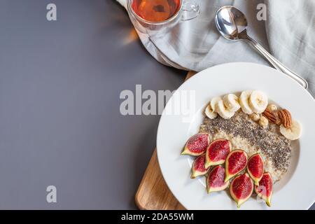 un bol avec petit déjeuner porridge de flocons d'avoine avec des graines de chia super-alimentaire, figues fraîches et banane sur une planche de bois sur fond gris, servi avec thé noir, grené d'en haut Banque D'Images