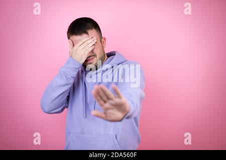 Jeune homme beau portant un sweat-shirt décontracté sur fond rose isolé couvrir les yeux avec les mains et faire un geste d'arrêt avec triste et la peur de l'expressio Banque D'Images