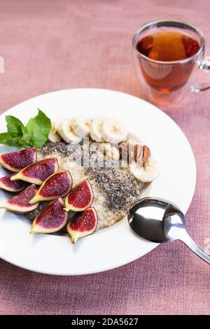 un bol avec porridge de flocons d'avoine pour le petit-déjeuner avec graines de chia super-alimentaire, figues fraîches et banane sur fond rose, servi avec du thé noir Banque D'Images