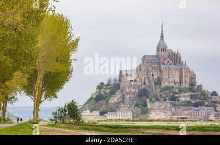 Deux personnes marchent le long d'un chemin bordé d'arbres avec la célèbre abbaye française du Mont St Michel au loin. Banque D'Images