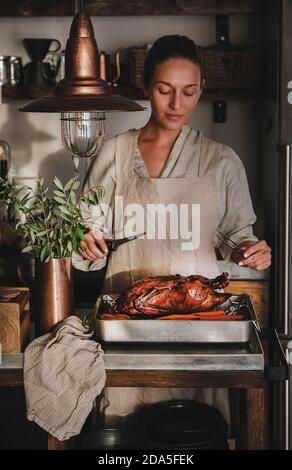 Jeune femme en tablier cuisant et sculptant du canard rôti au four entier pour les fêtes de Thanksgiving ou la fête de Noël dans l'intérieur de la cuisine. Traditionnel automne ou hiver vacances confort concept alimentaire Banque D'Images