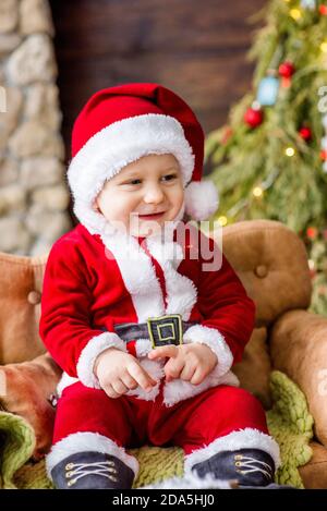 Un petit enfant en costume rouge du Père Noël, assis sur le canapé de la résidence, tenant un cadeau entre ses mains. Carte du nouvel an. Le garçon blond rit. Banque D'Images