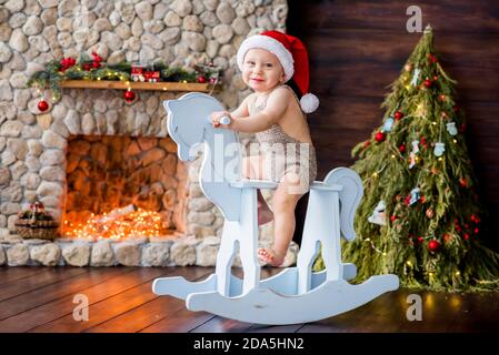 Un petit garçon manne un cheval en bois vintage près d'un arbre de Noël avec guirlande de lumières, une cheminée en pierre. L'enfant est vêtu d'une barboteuse tricotée Banque D'Images