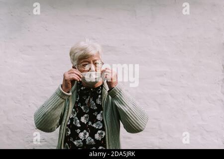 La femme caucasienne âgée avec des lunettes met le masque de visage à l'avant De la paroi blanche pendant la quarantaine Covid - 19 coronavirus Banque D'Images
