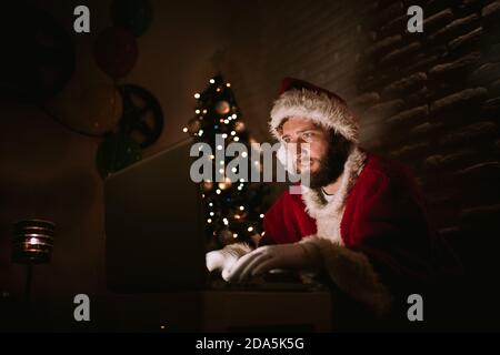 Un jeune caucasien habillé comme le Père Noël est assis dans une pièce à côté de l'arbre de Noël et regarde un ordinateur portable tout en parlant sur un appel vidéo. Banque D'Images