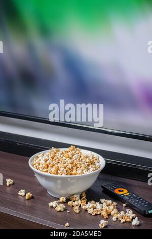 Un bol blanc rempli de pop-corn et un peu de pop-corn sur la table, devant un téléviseur LED avec télécommande. Banque D'Images