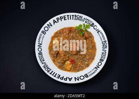 Délicieux ragoût maison de lentilles, de tomates, de chorizo espagnol, de viande et de pommes de terre servi dans un plat profond avec un motif noir sur une surface noire. Banque D'Images