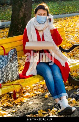 Femme de 50 ans et plus avec masque médical de protection assise sur un banc jaune dans le parc d'automne et parlant par téléphone avec une boîte pour pizza. Banque D'Images