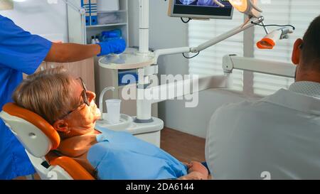 Vieille femme chez le dentiste ayant un traitement dentaire dans le bureau d'implantologie. Orthodontiste éclairant la lampe et parlant au patient assis sur une chaise stomatologique pendant que l'infirmière prépare les outils pour la chirurgie. Banque D'Images